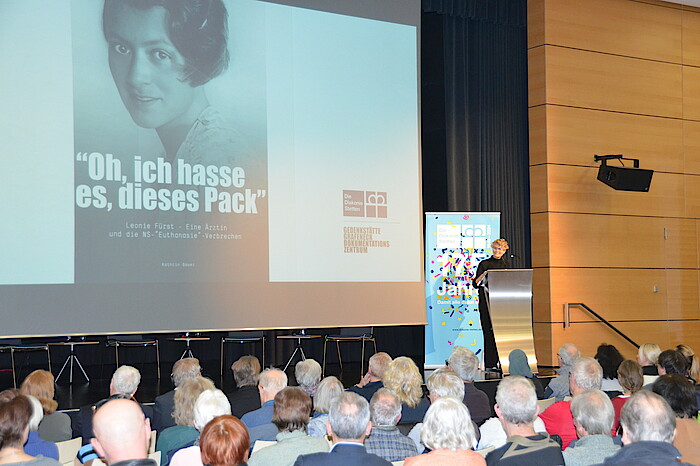 Referentin steht auf der Bühne vor Publikum, auf der Leinwand ein historisches Foto von Leonie Fürst und der Satz "Oh, ich hasse dieses Pack"