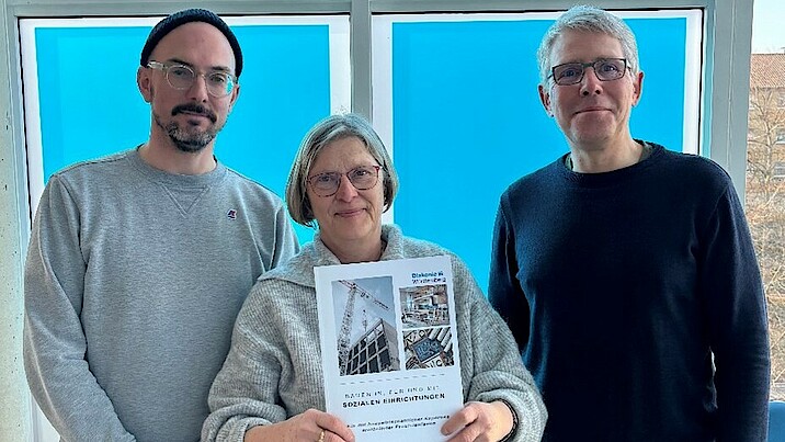 Eine Frau und zwei Männer stehen vor einem blauen Fenster. Die Frau hält eine Publikation in der Hand. 