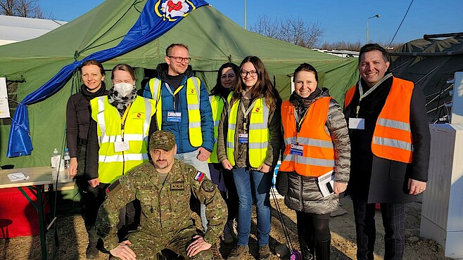 Acht Personen sitzen vor einem Zelt, das sich an der ukrainischen Grenze befindet, in der Sonne und lachen in die Kamera