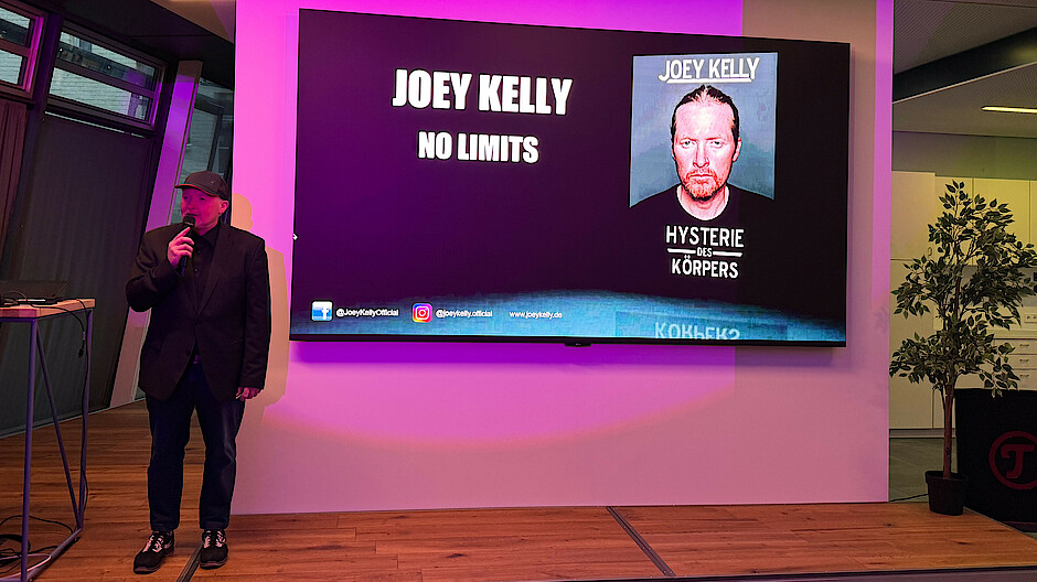 Joey Kelly steht und spricht vor einer Leinwand, auf der "no limits" steht.