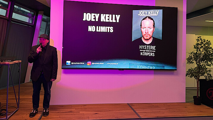 Joey Kelly steht und spricht vor einer Leinwand, auf der "no limits" steht.