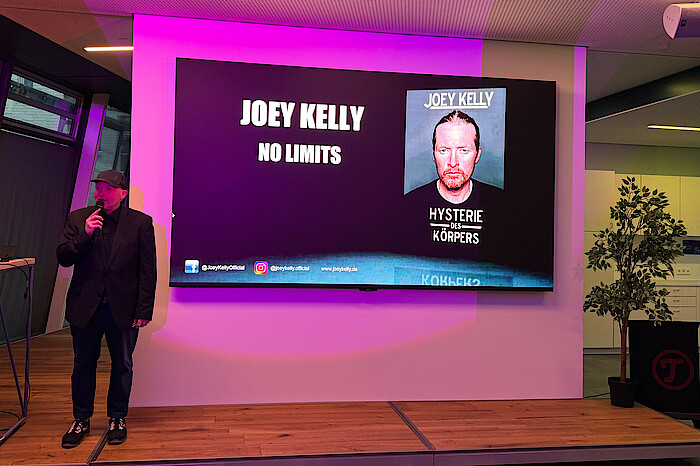 Joey Kelly steht und spricht vor einer Leinwand, auf der "no limits" steht.