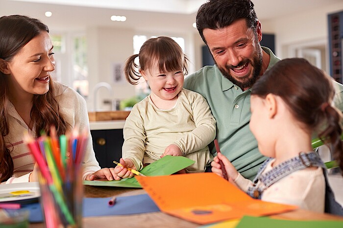 Mutter, Vater und zwei Kinder, davon ein Mädchen mit Down-Syndrom, sitzen an einem Tisch und malen.