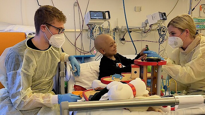 Bild zeigt zwei Helfer die im Krankenhaus an einem Bett sitzen und mit dem kranken Kind spielen